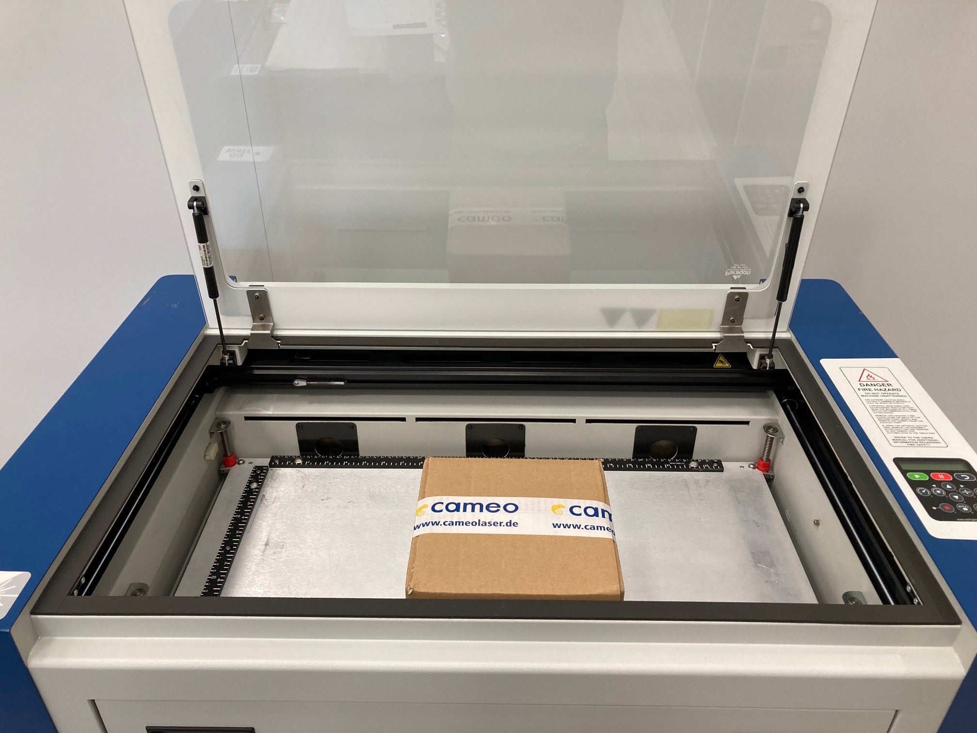 Geöffnete Epilog Zing 24 Maschine mit Schneidbrett, Paket, Glashaube und Einstellungsmenu.